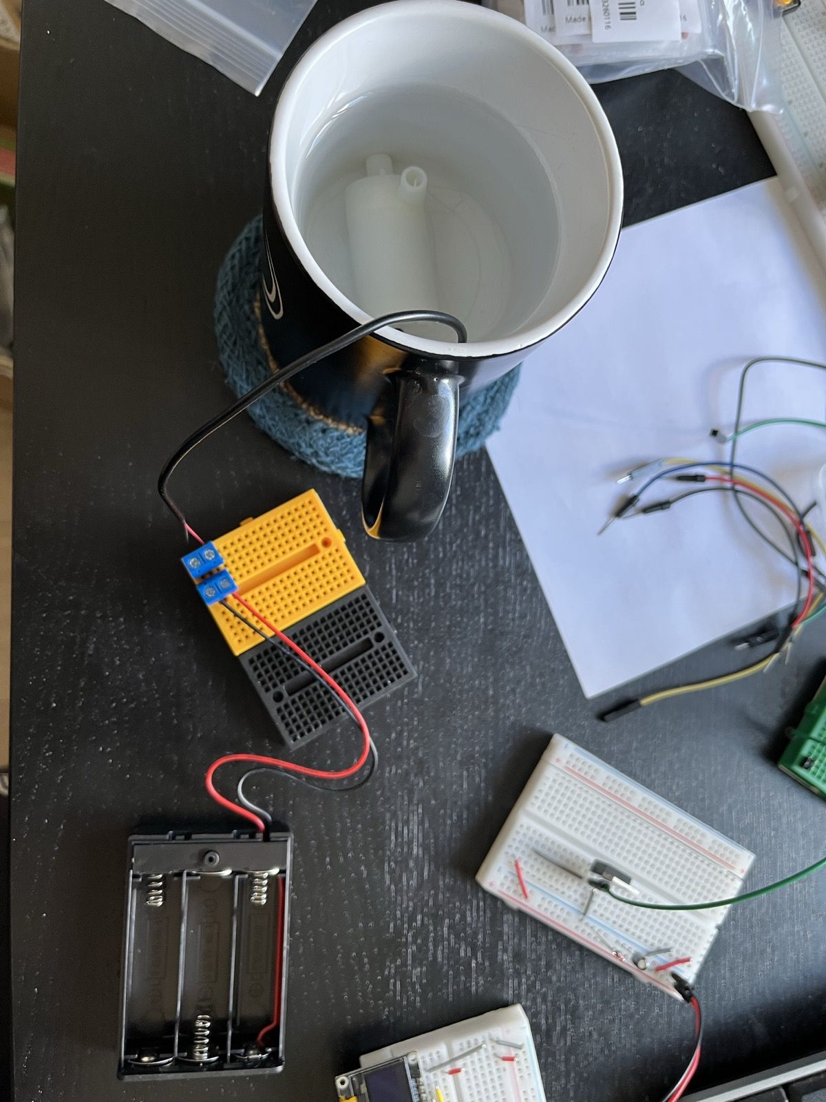 An amphibious water pump, submerged in water inside a black mug. A wire extends out of the mug and connects to an empty battery pack, via a yellow and black breadboard. Scattered around the components is the usual detritus you'll find on my desk most days.